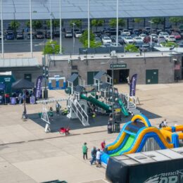 Outdoor event at a plaza with vendor tents, a science fair-like setup, and a large colorful inflatable slide in the foreground, and a parking lot with cars in the background under a solar canopy.